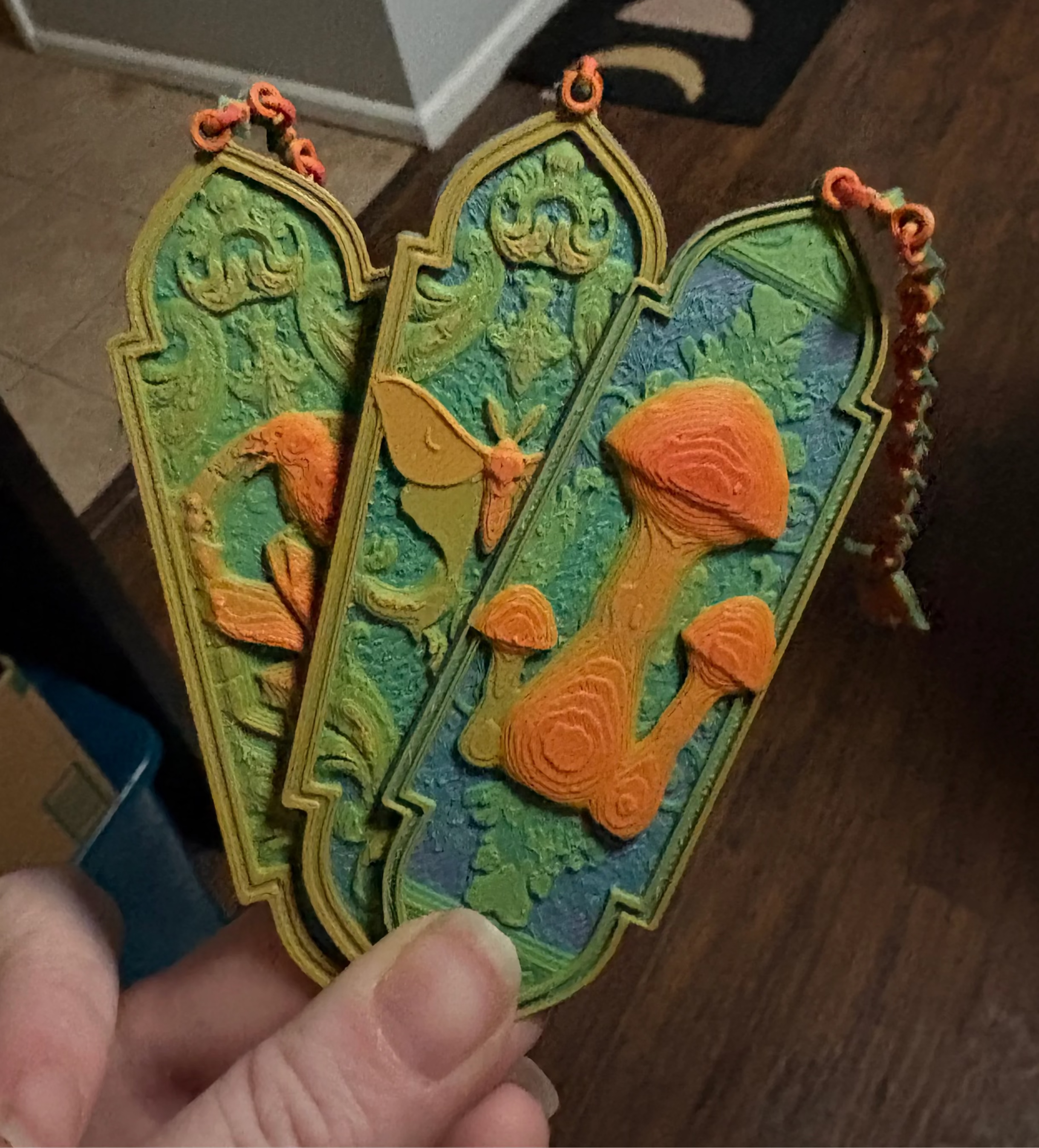 A hand holds three bookmarks that feature a raven, luna moth, and mushrooms. They are neon in color, fading through a rainbow with the lowest layers being blue and green and the upper layers orange. 
